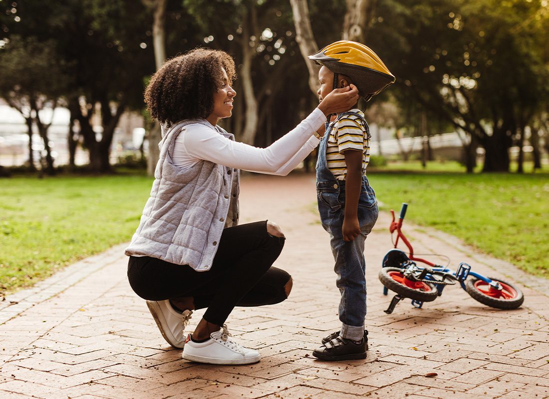 Group Voluntary Supplemental Life Insurance - Mother Placing a Protective Helmet on Her Son Before Riding a Bike in the Park on a Sunny Day