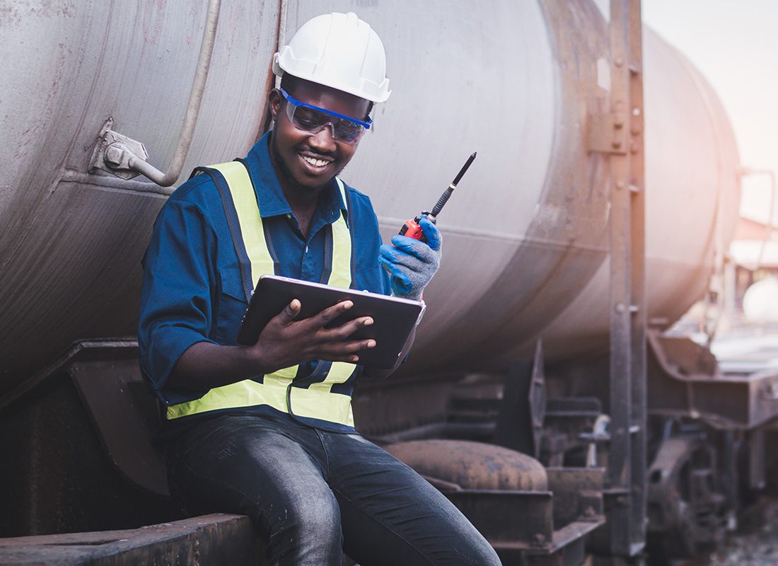 Contact - Portrait of a Cheerful Young Engineer Holding a Tablet and Handheld Radio in his Hands as he Sits Next to a Train in Need of Repairs
