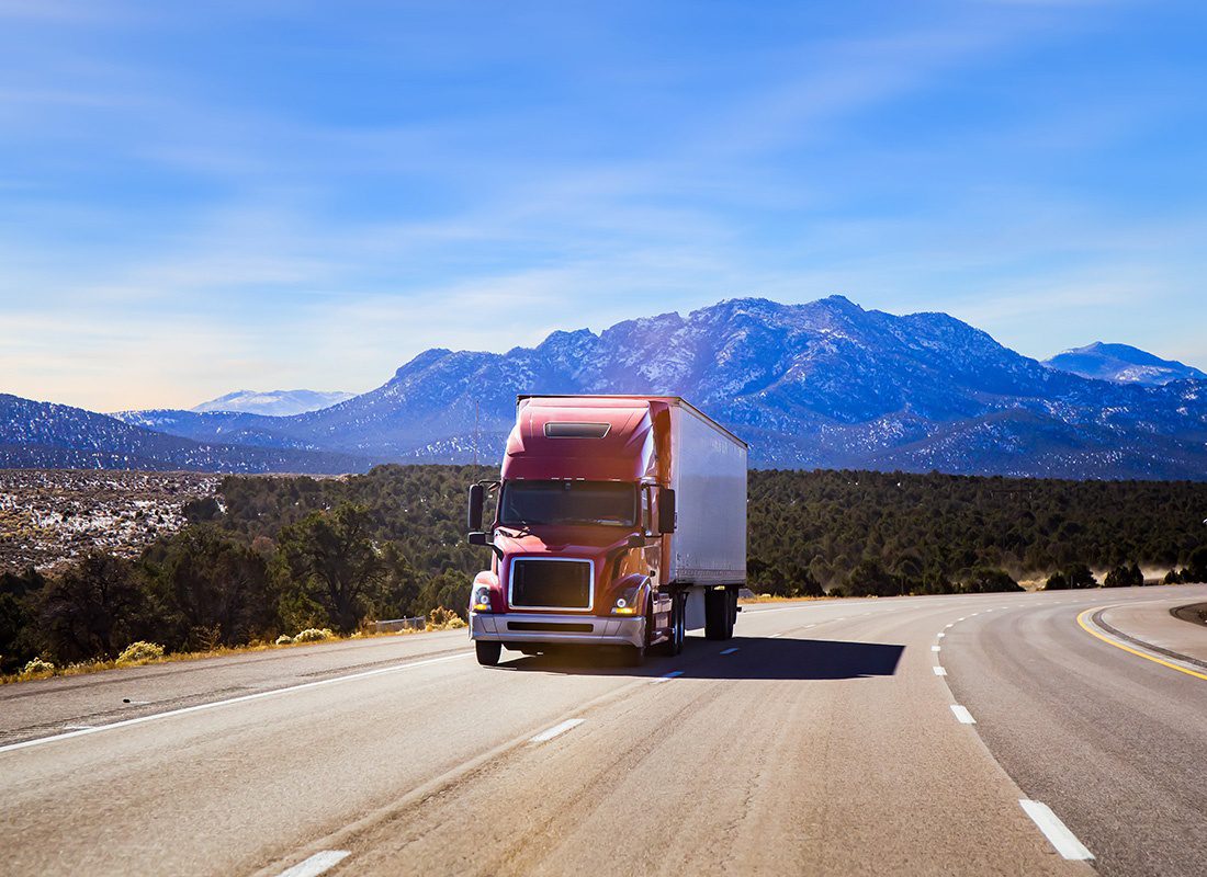 Insurance by Industry - View of a Red Truck with a Trailer Driving on an Empty Highway with Scenic Views of the Mountains Against a Blue Sky in the Background